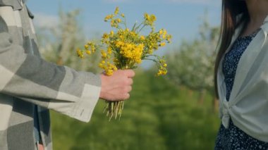 Güneşli bir günde bir buket sarı kır çiçeğiyle el ele tutuşan genç bir çiftin yakın çekimi. Sevgi ve sıcaklıkla dolu yemyeşil bir çayırda yumuşak romantik bir ortam..