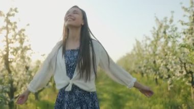Gülümseyen genç bir kadın, çiçek açan ağaçlar ve yeşil çimlerle dolu, ılık esintinin ve huzurlu kırsal atmosferin tadını çıkararak güneşli bir meyve bahçesinde geziniyor..