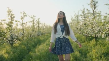 Mutlu genç bir kadın, çiçeklerle ve yeşilliklerle çevrili bahar çiçekleriyle dolu bir bahçede yürüyor. Yüzünde güneşi ve saçlarındaki meltemi hissediyor..