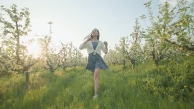 Neşeli genç bir kadın, güneşli bir bahar gününde çiçek açan ağaçların bahçesinde mutlu bir şekilde yürüyor, kırsal kesimin doğal güzelliği ve temiz havasıyla ıslanıyor..