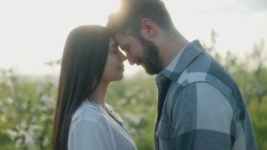 Genç bir çift, tomurcuklanan bir meyve bahçesinde, alınları birbirine değen yakın duruyor. Arkalarında güneş ışığı parıldar, sıcak ve yumuşak bir atmosfer yaratır. İlkbahar çiçeklerini sarar.