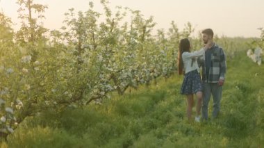 Çiçekler ve yemyeşil çiçeklerle çevrili, el ele güzel bir meyve bahçesinde birkaç gezinti. Akşamın erken saatlerindeki yumuşak ışık romantik bir atmosfer yaratır..
