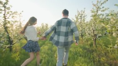 Gülen bir çiftin tam çiçeklerle dolu bir bahçede koşuşunun, güneşin tadını çıkarışının, bereketli manzarasının ve bahar akşamının taze büyüsünün arka görüntüsü..