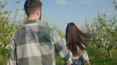 Mutlu genç çift dışarıda birlikte vakit geçiriyor, kadın erkeğin elini çekiyor, güneşli bahar zamanı yeşil bir bahçede neşeyle gülümsüyor..