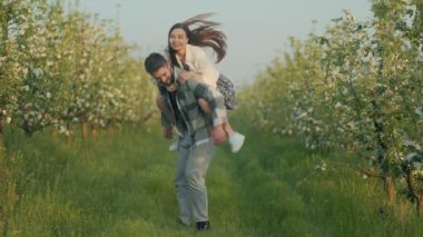 Güzel bir meyve bahçesinde erkek arkadaşını sırtında taşıyan neşeli genç bir kadın. Taze yeşil çimenler ve ılık bahar güneşi altında çiçek açan ağaçlarla çevrili..