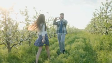Genç bir çiftin gün batımında çiçek açan bir meyve bahçesinde neşeli bir anın keyfini çıkardığı romantik bir sahne, yemyeşil ve ılık güneş ışığında çiçek açan ağaçlarla çevrili..