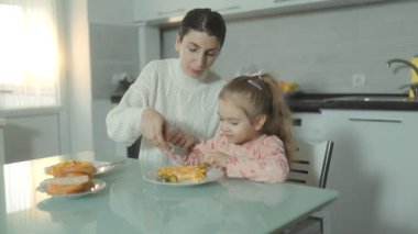 Bir anne ve küçük kızı mutfak masasında oturmuş sıcak ve aile ortamında birlikte yemek yiyorlar ve kalıcı anılar yaratıyorlar..