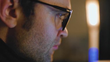 Düşük ışıklandırmalı bir ortamda oturan, gözlük takan ve bir işle uğraşırken güçlü bir odak noktası gösteren bir adam. Ortam sıcak bir aydınlatma ve kararlılık hissi içeriyor..