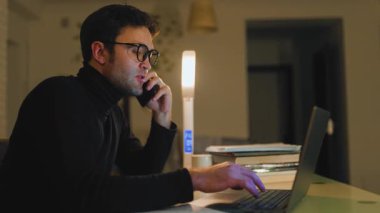 Siyah boğazlı kazak giyen bir adam masada oturuyor, telefonla konuşurken dizüstü bilgisayarına odaklanıyor..