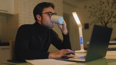 Genç bir adam sıcak bir atmosferde bir masada oturuyor, bilgisayarına odaklanırken kahvesini yudumluyor. Sahne, gece verimliliğinin ve yaratıcılığının özünü yakalıyor..