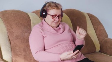 Yaşlı bir kadın rahat bir kanepede, akıllı telefon kullanarak ve kulaklık takarak vakit geçirmeyi seviyor. Sıcak bir iç mekanda dinlenirken en sevdiği melodilerle derinden ilgilenir..