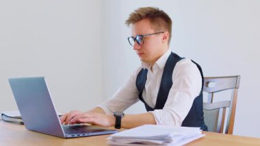 Resmi kıyafet giymiş genç bir adam dikkatle bir masada oturur ve dizüstü bilgisayarda yazar. İyi aydınlatılmış ofiste asgari dekorasyon ve yakınlarda bir yığın kağıt bulunuyor..