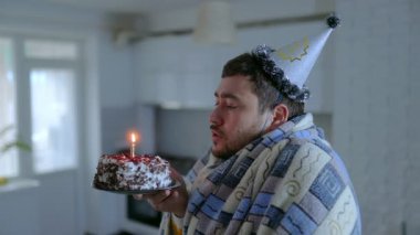 Üzgün, sakallı beyaz bir adam, battaniyeye sarılmış, soğuk bir evde oturup doğum günü pastasına mum üflüyor. Sahne yalnızlığı ve yalnızlığı yansıtıyor..