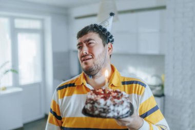 Sarı tişörtlü sakallı bir adam doğum gününü yalnız kutluyor, elinde yanan bir pasta var ve mutfakta parti şapkası takarken hayal kırıklığına uğruyor..