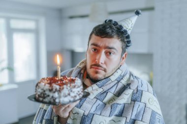 Soğukla savaşmak için battaniyeye sarılmış orta yaşlı bir adam, mutfağında yanan bir doğum günü pastası tutuyor, yalnız kutlamasında üzgün ve yalnız hissediyor..