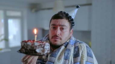 Sakallı üzgün bir adam soğuk bir mutfakta oturuyor, battaniyeye sarılı, yanan bir pasta tutuyor, yalnızlığa ve rahatsızlığa katlanırken gözle görülür bir şekilde üzgün..