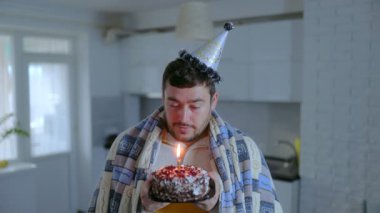 Yalnız ve soğuk bir kutlama, parti şapkalı bir adamı, battaniyeye sarılı bir doğum günü pastası tutarken, üzüntüsünü ve hayal kırıklığını yansıtır..