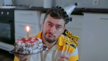 Orta yaşlı, parti şapkalı bir adamın elinde mumla doğum günü pastası tutarken, yüzünde kasvetli bir ifadeyle mutfakta tek başına oturduğu duygusal bir sahne..