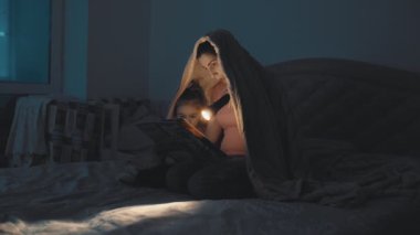 Bir battaniyenin altında, bir anne ve kız bir kitabın üzerine bağlanır, sayfaları aydınlatmak için bir el feneri kullanır, huzurlu bir gece anısı yaratır..