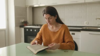 Genç bir kadın mutfak masasında oturmuş İncil okuyor, inancını çalışıyor ve evinin huzurlu atmosferine sakin bir şekilde yansıyor..