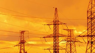 Enerji akışını ve ızgara karmaşıklığını yansıtan canlı turuncu günbatımına karşı siluetli çoklu elektrik direklerinin ve yoğun elektrik hatlarının hızlandırılmış görüntüsü.