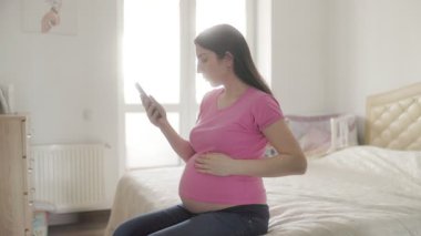 Hamile genç bir kadın yatak odasında rahat bir şekilde oturuyor, interneti gezmek için cep telefonunu kullanıyor. Çevrimiçi olurken elini nazikçe karnına koyar..