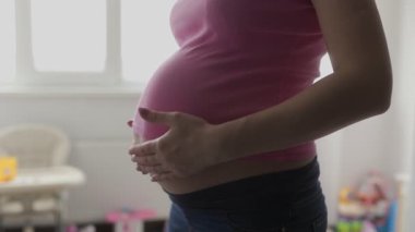 Pembe bluz ve kot giyen bir kadın hamile karnını güzelce aydınlatılmış bir odada sarkıtır..