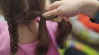 Bir insan ustalıkla bir çocuk saçını örüyor, düzgün kıvrımlara ve güvenli kravatlara odaklanıyor. Sıcak atmosfer, ev ortamında neşeli ve sevgi dolu bir bağ deneyimi olduğunu gösteriyor..