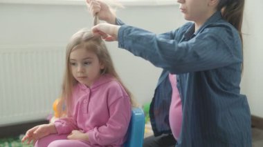 Bir anne, genç kızının saçlarını hoş, renkli bir ortamda nazikçe şekillendirir. Kız sakince oturur, pembe bir kıyafet giyer, oyuncaklar etrafa saçılırken..