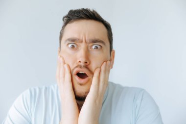 Açık renkli bir odada ellerini yüzüne koyan bir adam şok olmuş bir ifade sergiliyor. Onun geniş gözleri ve açık ağzı güçlü bir şaşkınlık ya da inanmazlık duygusu iletir..