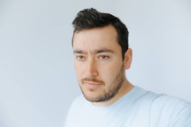 Açık renk sakallı ve koyu renk saçlı bir adam kameraya bakıyor. Açık mavi bir gömlek giyiyor ve arka planı yumuşak, nötr bir renk..