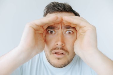 Bir adam ellerini yüzünü çerçevelemek için kullanırken geniş gözleri ve çatık kaşlarıyla şaşkınlığını ifade eder. Çevre aydınlık ve nötr. Sürprizi artırıyor..