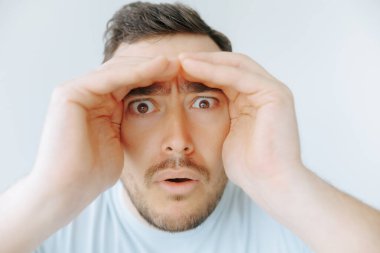 Şok olmuş bir yüz ifadesi olan bir adam ellerini alnının üstüne koyarak dikkatle ileriye bakar. Şaşırmış görünüyor, bir anlık merakı ya da güvensizliği vurguluyor..