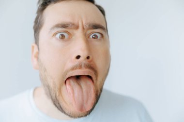 Bir adam, dilini uzatarak ve kaşlarını kaldırarak abartılı bir ifade sergiliyor. İyi ışıklandırılmış bir odada açık renkli bir arkaplanda eğleniyor ya da şaşırıyor gibi görünüyor..