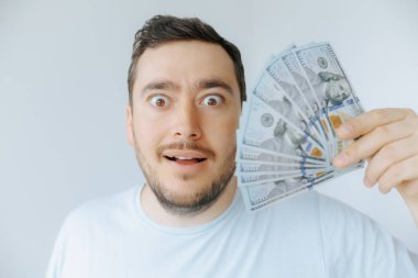 Şaşırmış bir yüz ifadesi olan bir adam sağ elinde bir tomar para tutar. Açık renkli bir duvarın karşısında durmuş mali durumuyla ilgili heyecan ve mutluluğu aktarıyor..