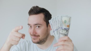 Kısa siyah saçlı bir adam kamerayı işaret ediyor ve içi para dolu, minimalist bir odada temiz bir kap tutuyor. İfadesi çekici ve eğlenceli..