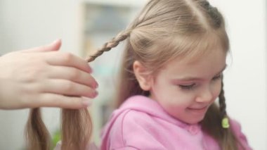 Uzun saçlı genç bir kız, bakıcı gibi gülümsüyor ve saçlarını kapalı bir ortamda nazikçe örüyor. Atmosfer neşeli ve besleyici, özel bir bağ kurma anını vurguluyor..