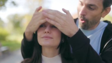 Genç bir çift spontane bir anın tadını çıkarırken, erkek, partnerinin gözlerini arkasından şakayla örtüyor. Kahkahaları sonbahar parkı ayarlarını dolduruyor, bağlarını yansıtıyor..