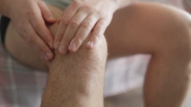 Bir adamın diz eklemlerine krem sürerken, ağrı kesici ya da cilt bakımı için nazikçe ovalarken, doğal ışık alan bir ev ortamında.