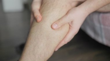 Bir adamın alt bacağını iki eliyle ovalarken, muhtemelen ağrı, yorgunluk ya da rahatsızlıktan dolayı. Görüntü, deri dokusu ve vücut kıllarının detaylarını gösteriyor..