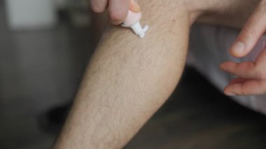 Ağrı kesici veya cilt bakımı için bacağına beyaz krem süren ve yayan bir adamın yakın plan görüntüsü, el hareketlerini ve detaylarını kapalı alanda rahat bir şekilde gösteriyor..