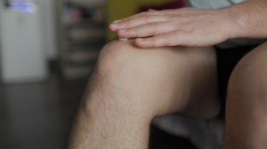 Bir eliyle ağrıyan dizine krem sürerken diğer eliyle bölgeyi ovuşturan bir adamın yakın çekimi. İç mekan ayarları eklem ağrısı veya rahatsızlık tedavisi öneriyor.