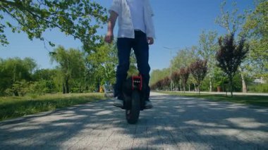 Neşeli genç bir adam güneşli bir günde yeşil bir şehir parkında elektrikli tek tekerlekli bisiklete biner. Mavi bir kask ve günlük kıyafetler giyerek, açık havada kaygısızca gezmekten hoşlanıyor..