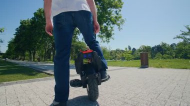 Şehir parkında elektrikli tek tekerlekli bisikletinin yanında duran kotlu ve kasklı sıradan bir genç göl kenarında güneşli bir günde bankta oturmaya hazırlanıyor..