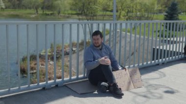 Evsiz bir adam, elinde bir karton levhayla beton kaldırımda oturuyor. İşsizlik ve evsizlikle mücadelesinin detaylarını yazıyor. Finansal zorlukların etkisini vurguluyor..