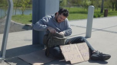 Şehir parkında, genç bir adam kaldırımda oturur ve küçük bir yemek yer. İşsizlik ve evsizlik yüzünden yaşadığı zorlukları ifade eden bir tabela tutuyor, zor gerçeklerle yüzleşiyor..