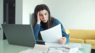 Evde çalışan stresli ve endişeli bir kadın, dizüstü bilgisayar kullanırken mali dökümanları ve faturaları analiz ediyor, tükenmişlik ve hayal kırıklığı belirtileri gösteriyor..