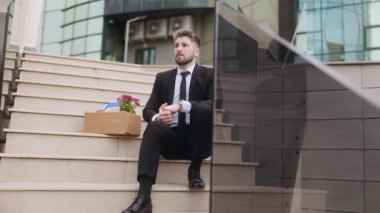 Kurumsal adam cam binanın önünde oturuyor. Yenilmiş bir görünüş ve kişisel kutusuyla. İş kaybını, işsizliği ve duygusal stresi simgeliyor..