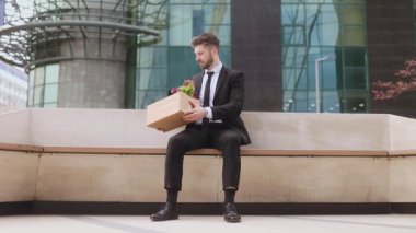 Beyaz yakalı bir işçi ofis binasının dışındaki bankta oturuyor, elinde kişisel eşyalar ve çiçeklerle dolu bir karton kutu var. Son işten çıkarılmasından dolayı derin düşüncelere dalmış ve üzgün görünüyor..
