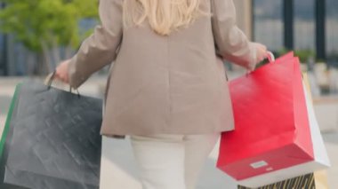 Kendine güvenen bej ceketli ve beyaz pantolonlu bir kadın ellerinde renkli alışveriş çantalarıyla dışarı çıkıyor. Alışveriş bölgesinde modern sokak modası..